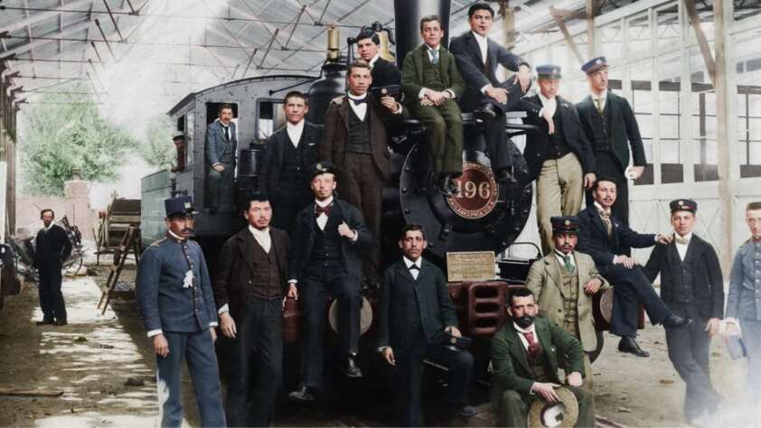 A group of men in late 19th-century clothing pose around and on a large steam locomotive inside a train shed, with some men in uniform and others in suits, standing or sitting casually on the engine.