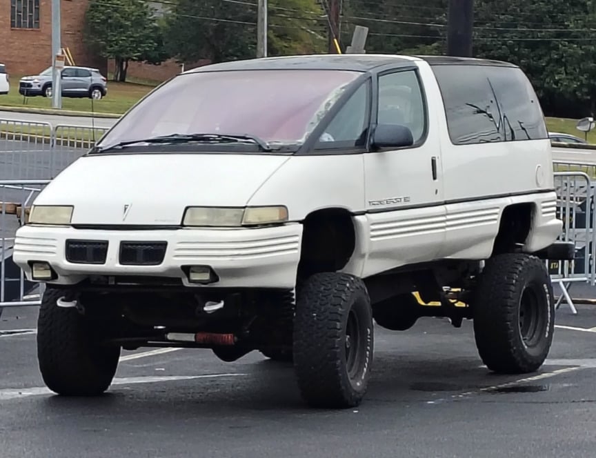 A white Pontiac Trans Sport minivan is modified with a lifted suspension and large off-road tires, parked outdoors on asphalt near metal barricades.