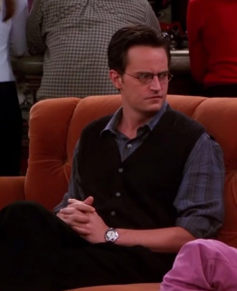 A man with short dark hair and glasses sits on an orange couch, wearing a dark vest over a blue plaid shirt, with his hands clasped and a serious expression, in a coffee shop setting.