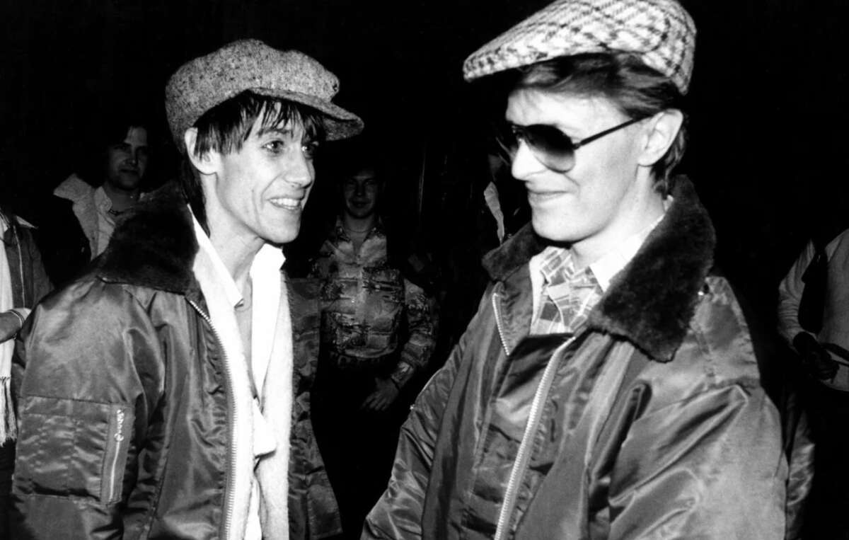 Two men wearing jackets and flat caps smile at each other. One wears sunglasses and a plaid cap, while the other has a light scarf. The scene appears candid and friendly, with people in the background. The photo is black and white.