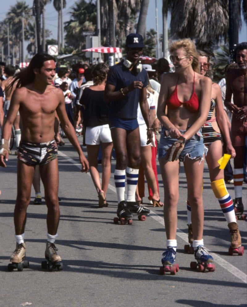 People roller skating on a sunny street lined with palm trees. The scene is lively, with men and women in swimsuits and retro clothing, enjoying the day. The atmosphere is relaxed and vibrant.