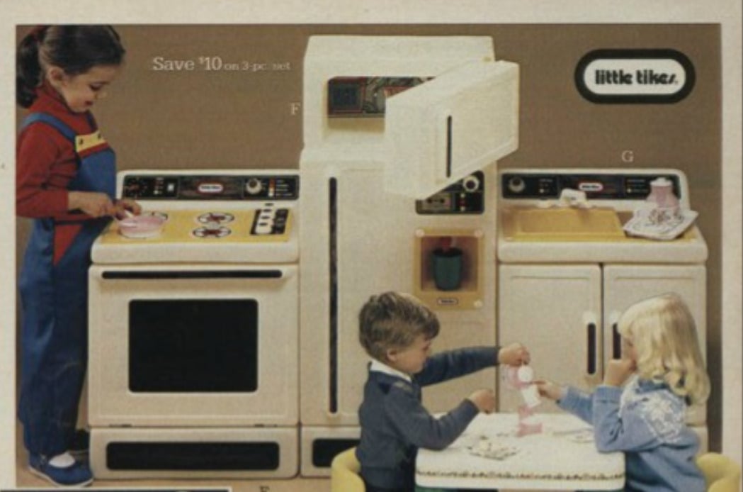 Three children play with a vintage Little Tikes toy kitchen set. One child pretends to cook at the stove, while two others sit at a small table, having a pretend tea party with toy dishes.