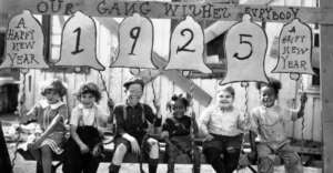 Six children sit on a wooden bench under a New Year banner reading &ldquo;OUR GANG WISHES EVERYBODY A HAPPY NEW YEAR 1925,&rdquo; smiling and holding large paper bells with the year 1925 and festive greetings.