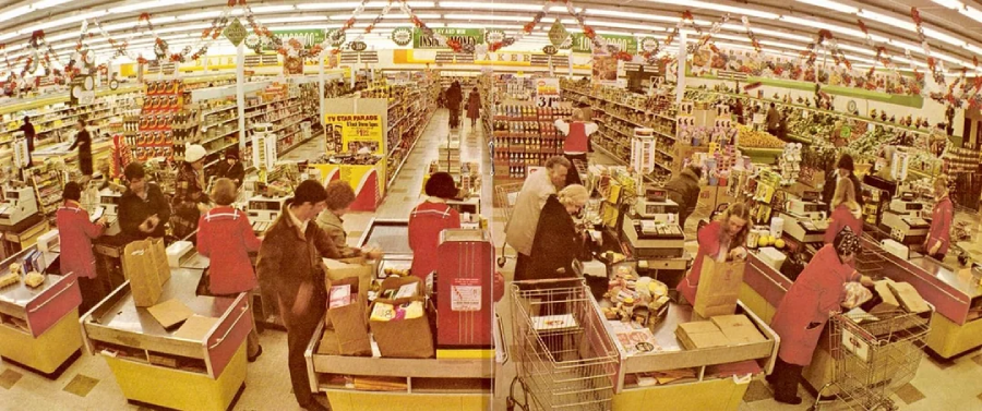 A busy supermarket in the 1970s with shoppers at checkout counters, cashiers in red uniforms, and aisles stocked with groceries. Holiday decorations hang from the ceiling, creating a festive atmosphere.