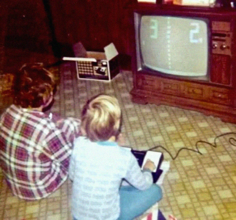 Two children sit on the floor in front of an old TV, playing a retro video game that resembles Pong. A game console and wires are visible, and the room has patterned linoleum flooring and wood-paneled walls.