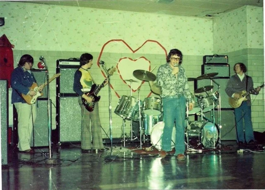 A vintage photo of a five-member rock band performing indoors. Three are playing guitars, one is on drums, and one sings into a microphone. A red heart is drawn on the wall behind them. The setting appears casual and retro.