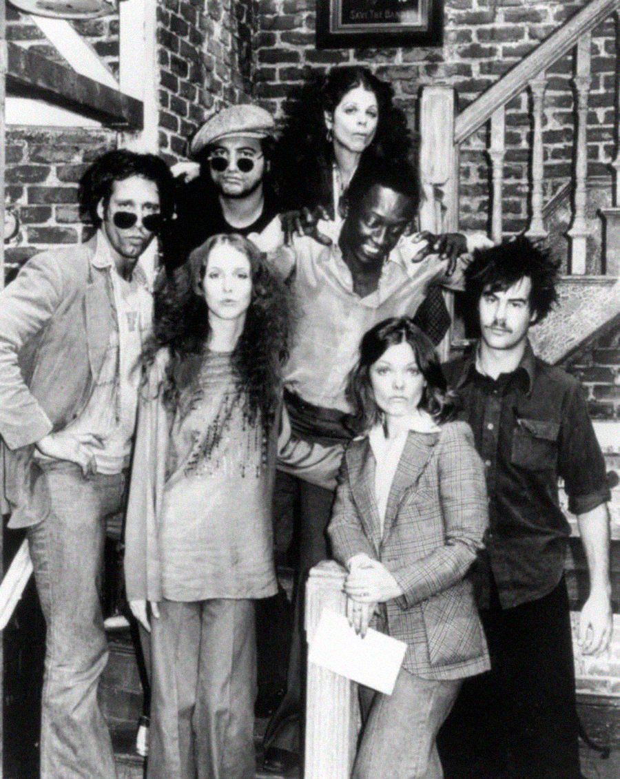 A black-and-white photo of seven people posing on a staircase in front of a brick wall. The group wears 1970s-style clothing, with a mix of casual and formal outfits and various facial expressions.