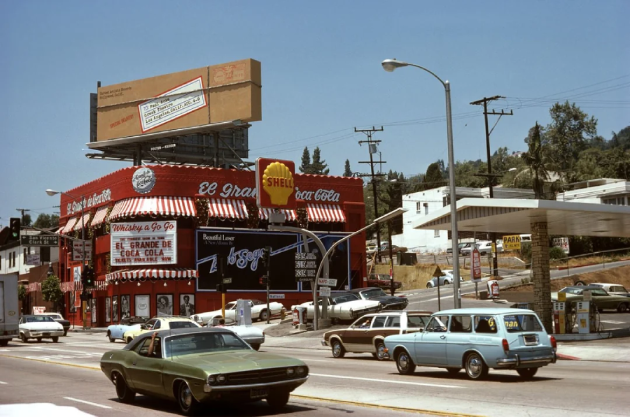 A busy 1970s street scene with vintage cars passing by El Drano, a red and white corner restaurant, and a Shell gas station. A large Coca-Cola billboard and palm trees are visible in the background.