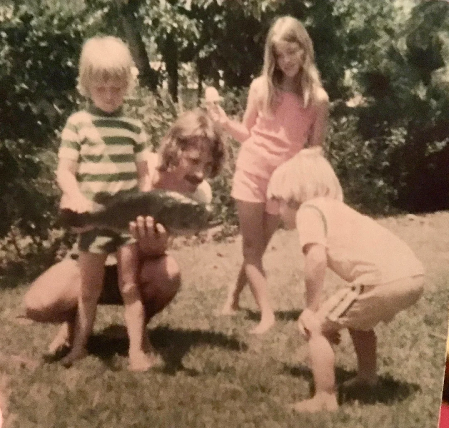 Three children and an adult outdoors on grass; the adult is kneeling and holding a large fish, while the children, barefoot and casual, look at the fish with curiosity. Trees and greenery are in the background.