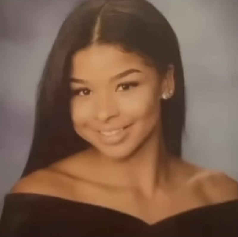 A young woman with long, straight dark hair smiles in a formal portrait, wearing an off-the-shoulder black top and stud earrings, posed against a softly blurred blue-gray background.