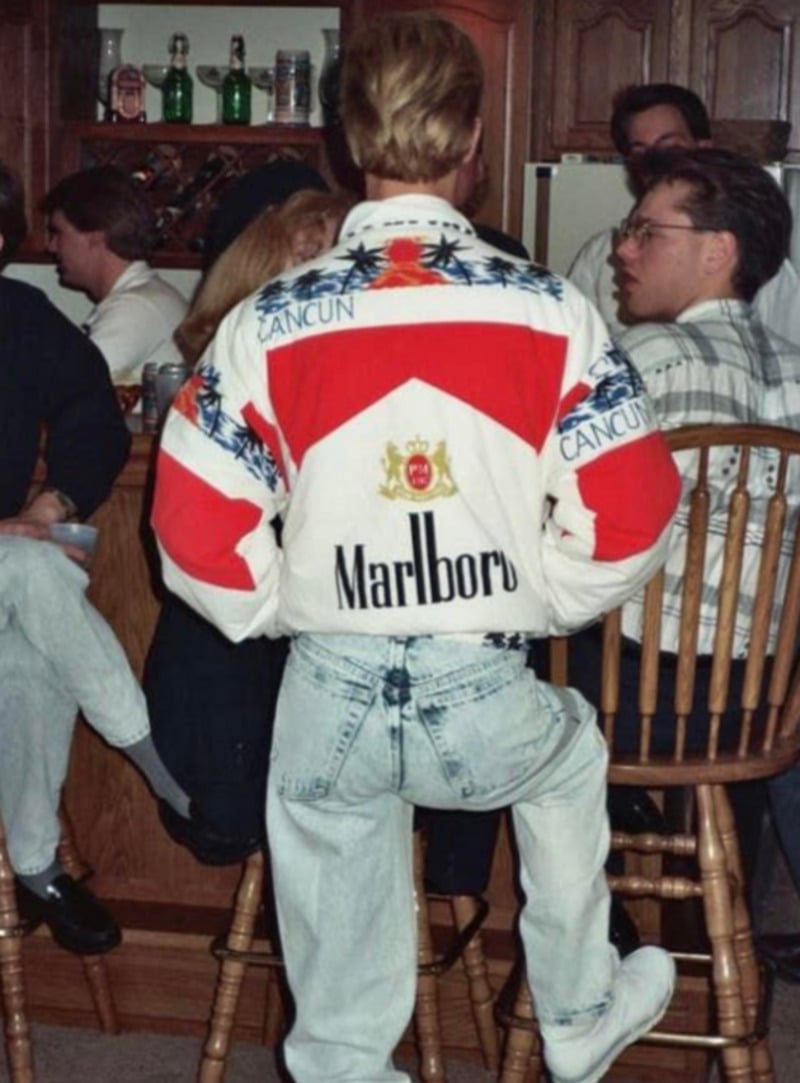 A person with blond hair stands with one leg on a wooden chair, wearing a colorful Marlboro Cancun jacket and light-washed jeans at a lively indoor gathering with several people and kitchen cabinets visible.