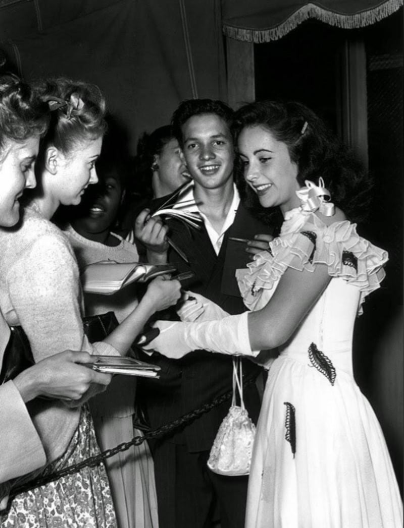 A young woman in an elegant dress and gloves smiles while signing autographs for a group of excited young fans, separated by a rope barrier. The photo is in black and white and appears to be from the mid-20th century.