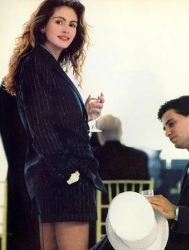 A woman in a pinstripe blazer and skirt smiles while holding a glass, standing beside a seated man who is looking down and holding a white hat. People and chairs are visible in the blurred background.