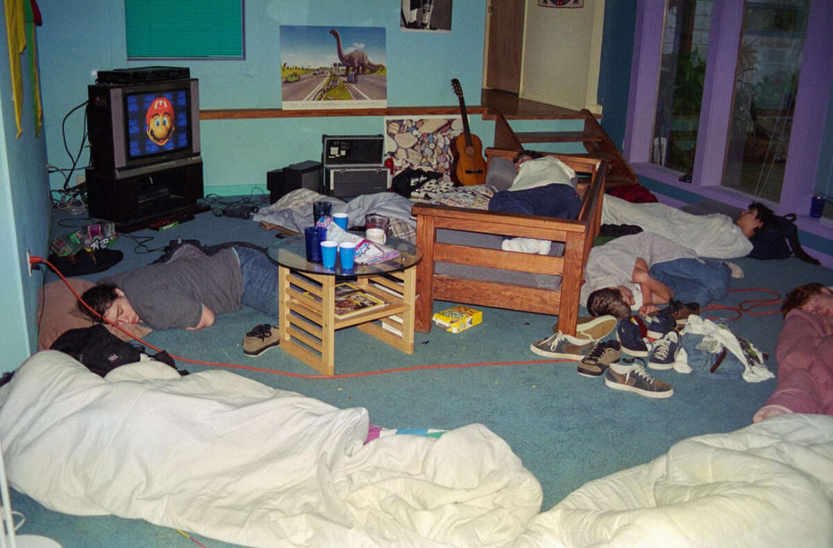 A group of people sleep on the floor and furniture in a messy living room with blankets, snacks, and cups scattered around. A TV shows a video game, and a guitar rests near the wall. Sunlight comes through large windows.