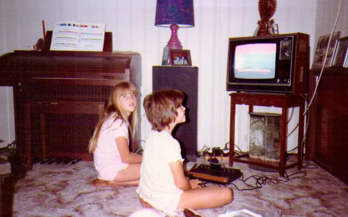 Two children sit on a carpeted floor playing a retro video game on a small TV. Behind them are a piano with sheet music and a lamp on a wooden table. The room has vintage decor and natural light from curtained windows.
