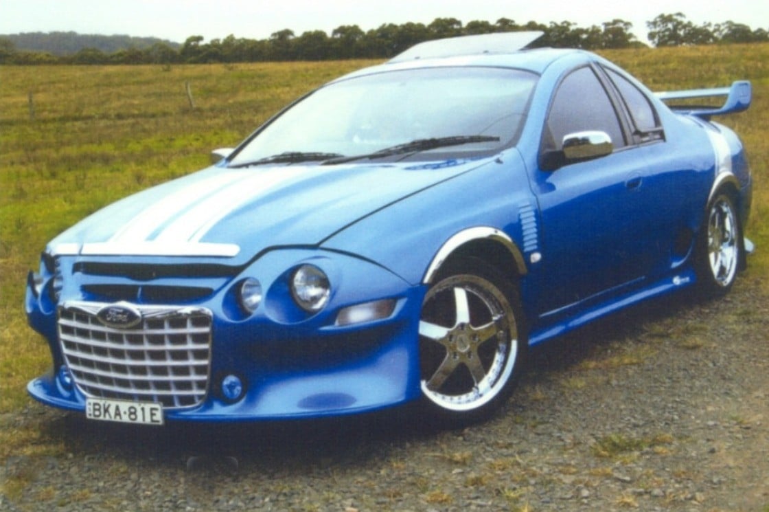 A bright blue modified car with a large rear spoiler, custom front bumper, silver racing stripes, and chrome rims parked on a gravel area near a grassy field.