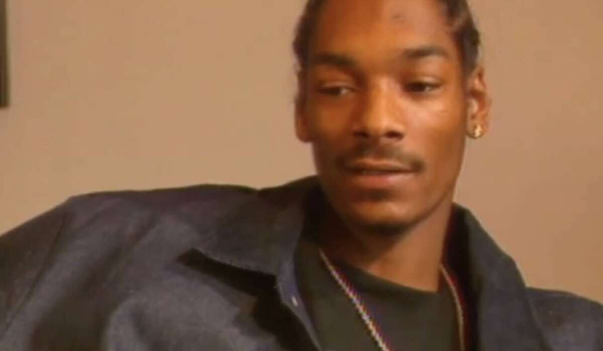 A man with braided hair and a goatee, wearing a dark jacket, black shirt, gold earring, and a chain necklace, sits and looks slightly to the side in a softly lit room.