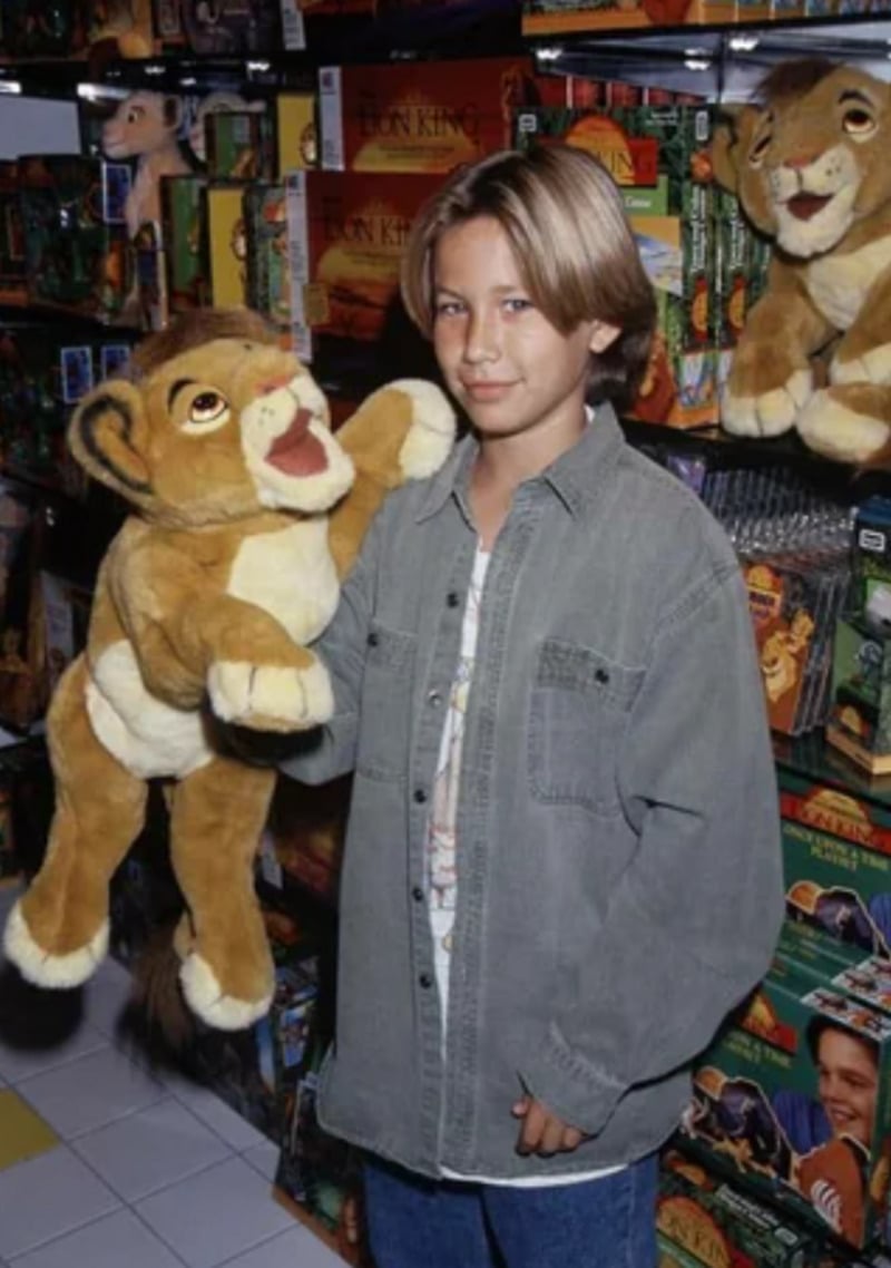 A young boy with blonde hair wearing a gray button-up shirt holds a large Lion King plush toy in a store filled with Lion King merchandise and toys.