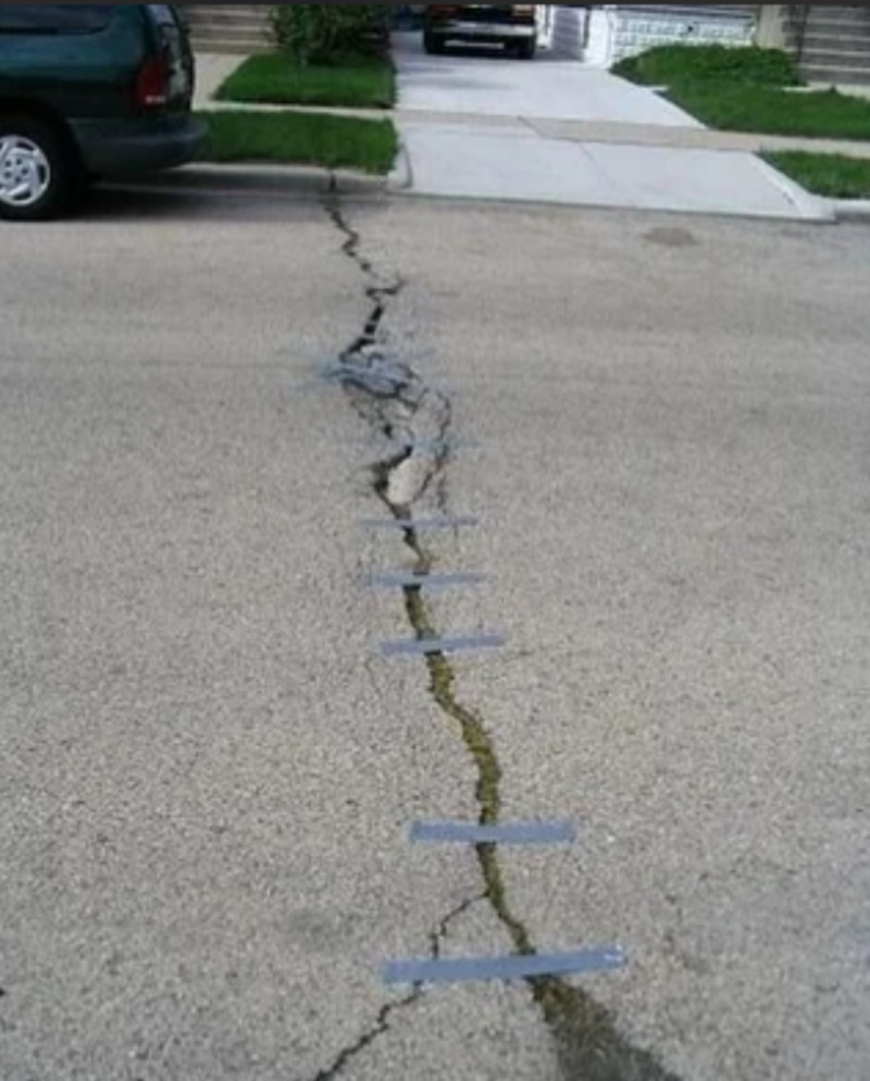 A cracked road with several strips of gray duct tape placed across the crack, attempting to hold the broken pavement together as a temporary fix. A car and driveways are visible in the background.