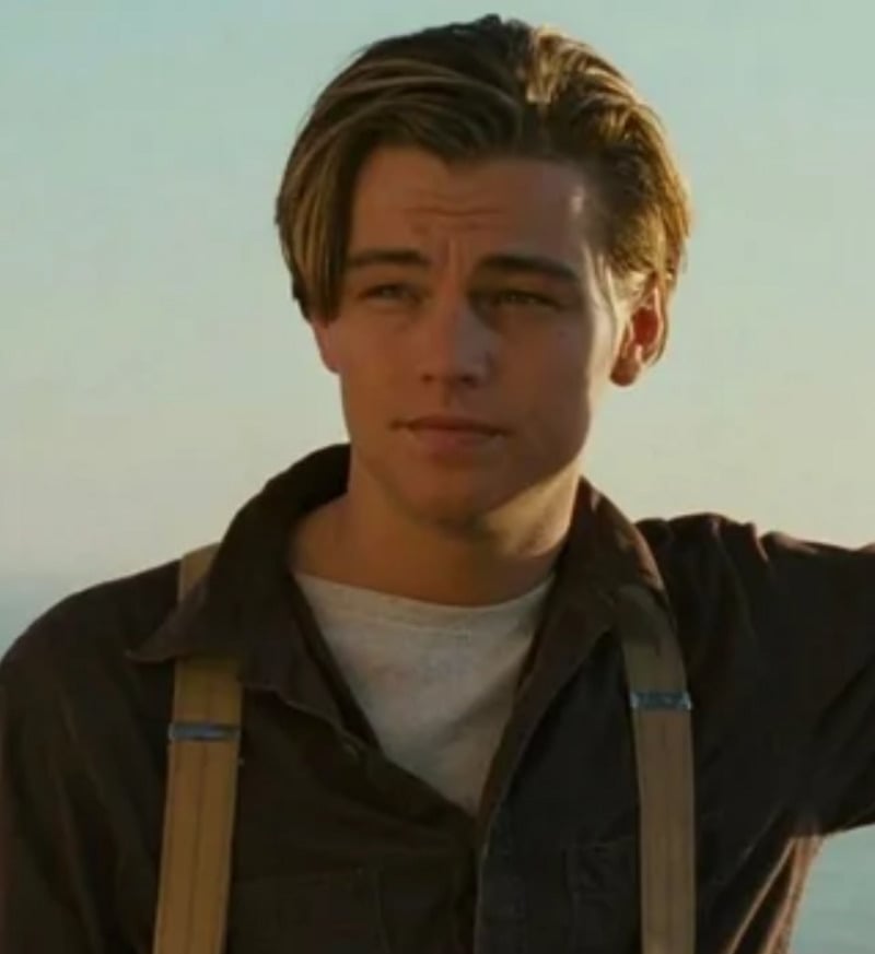 A young man with neatly styled brown hair, wearing a dark shirt over a white undershirt and tan suspenders, stands outdoors in natural daylight, looking thoughtfully into the distance.