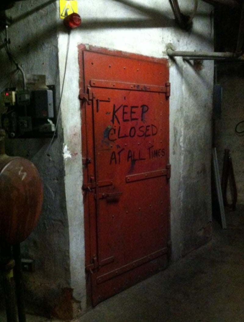 A heavy red metal door with large hinges is set in a concrete wall. The door has “KEEP CLOSED AT ALL TIMES” painted on it in black letters. The dimly lit area appears to be a basement or utility room.