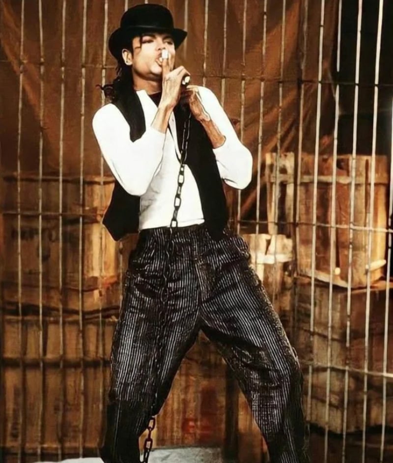 A man dressed in a black hat, black vest, and striped pants performs on stage, holding a microphone, with a chain around his neck. The background shows wooden crates and a wire fence.