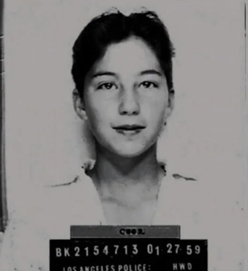 A black and white mugshot photo of a young woman with short dark hair, wearing a light-colored shirt. She faces the camera with a neutral expression. A police placard with numbers and text is visible at the bottom.