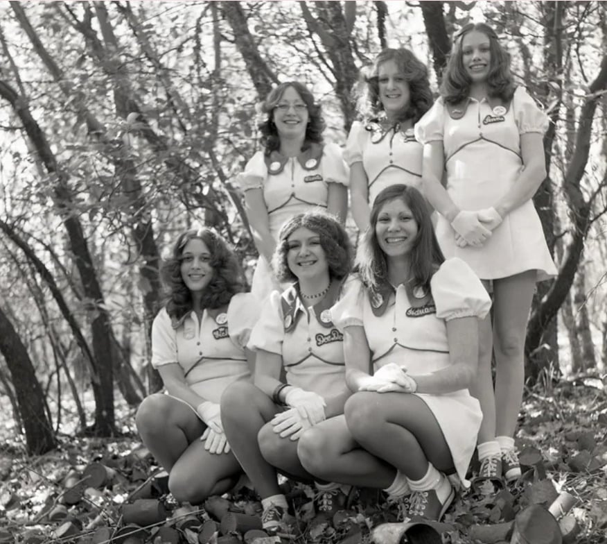 Six women in matching short-sleeve dresses, gloves, and headbands pose outdoors in a wooded area, smiling for the camera. The photo has a vintage, black-and-white look.