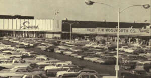 A busy parking lot filled with vintage cars in front of a shopping center featuring Sears, Zales, and F.W. Woolworth Co. stores. The image appears to be from the mid-20th century.