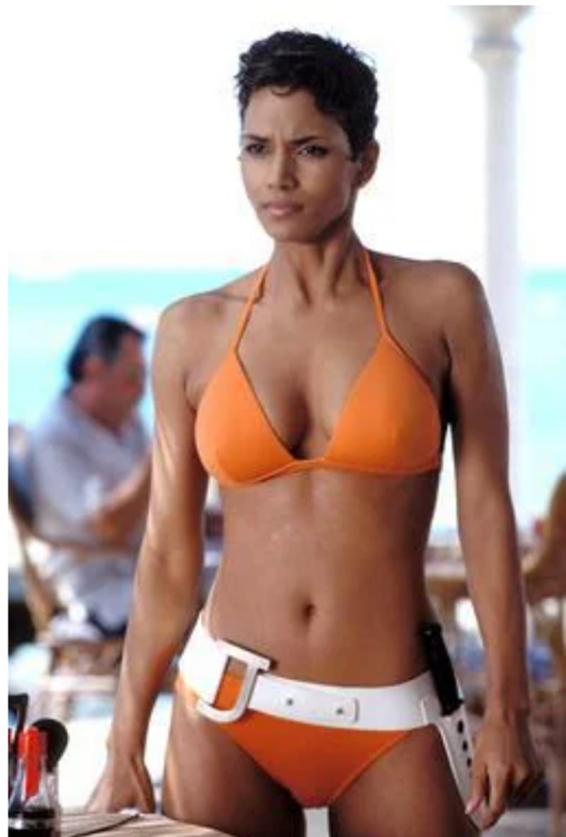 A woman with short hair wearing an orange bikini and a white belt stands confidently outdoors, with blurred people and the ocean in the background.