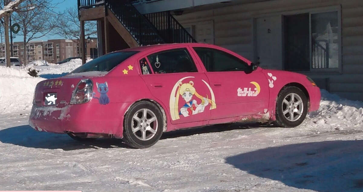 A pink car decorated with large Sailor Moon and Luna character decals, stars, and Japanese text is parked in a snowy lot next to a building on a sunny day.