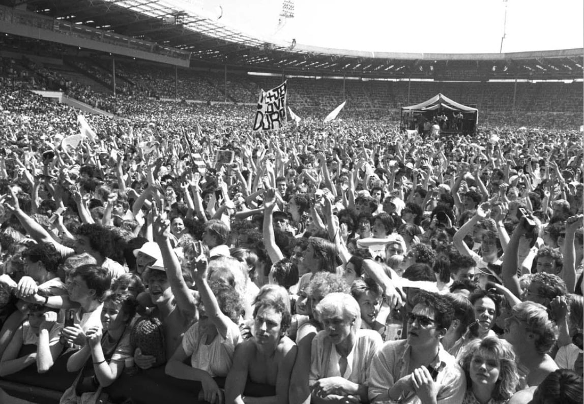A large, energetic crowd at an outdoor stadium concert, with many people raising their hands and cheering. Some hold signs and flags. The stage is visible in the distance under a canopy. The photo is in black and white.