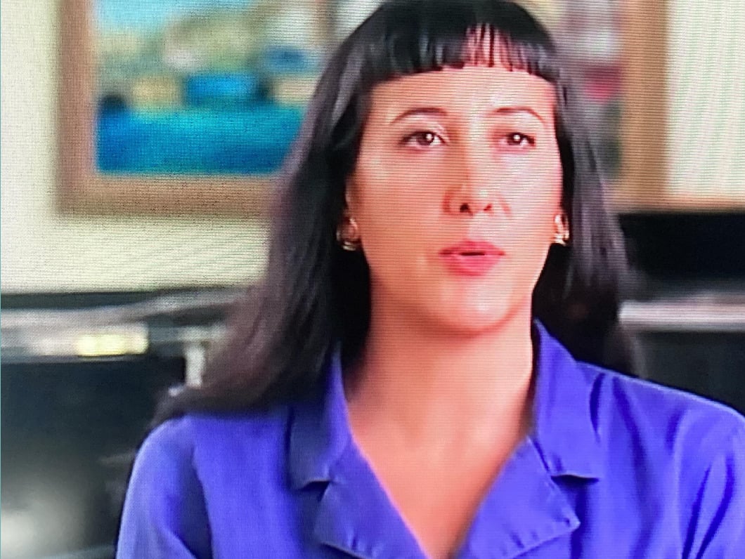 A woman with long dark hair and bangs, wearing a purple collared shirt, sits indoors with blurred paintings visible in the background.