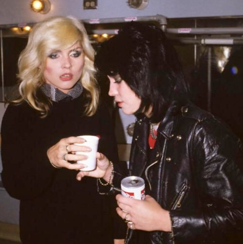 Two women with blonde and black hair stand together indoors. One wears a black sweater and scarf, holding a white cup, while the other wears a leather jacket and holds a can, both appearing to chat in a backstage setting.