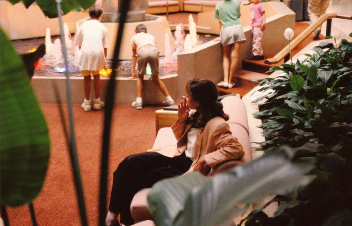 A woman sits on a couch surrounded by plants, looking away as children play at a colorful indoor fountain in the background.