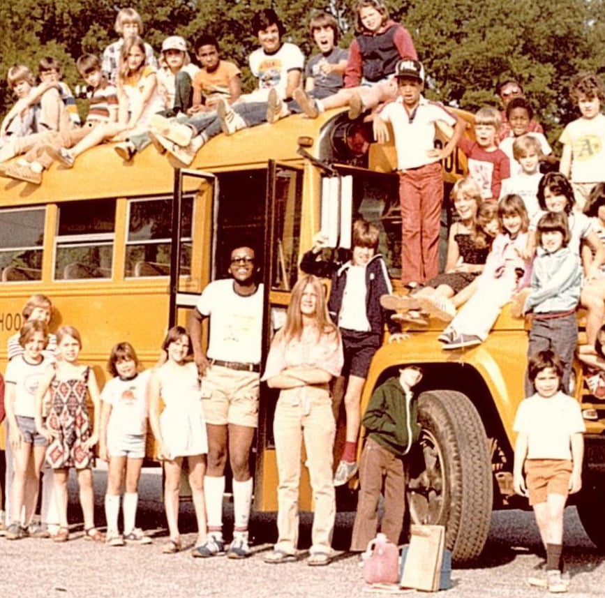 A large group of children and adults pose in and around a yellow school bus. Some sit on the roof, while others stand or sit in front. Trees are in the background, and everyone looks cheerful. The photo has a vintage feel.