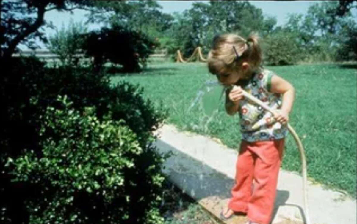 A young girl with pigtails, wearing a colorful top and red pants, drinks water from a garden hose while standing next to green bushes and grass on a sunny day.