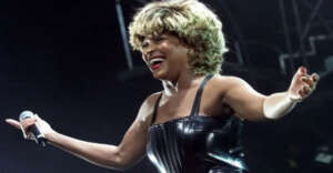 A woman with blond hair wearing a shiny black strapless outfit holds a microphone and smiles with her arms outstretched on stage under bright lights.