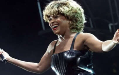 A woman with blond hair wearing a shiny black strapless outfit holds a microphone and smiles with her arms outstretched on stage under bright lights.