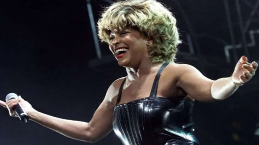 A woman with blond hair wearing a shiny black strapless outfit holds a microphone and smiles with her arms outstretched on stage under bright lights.