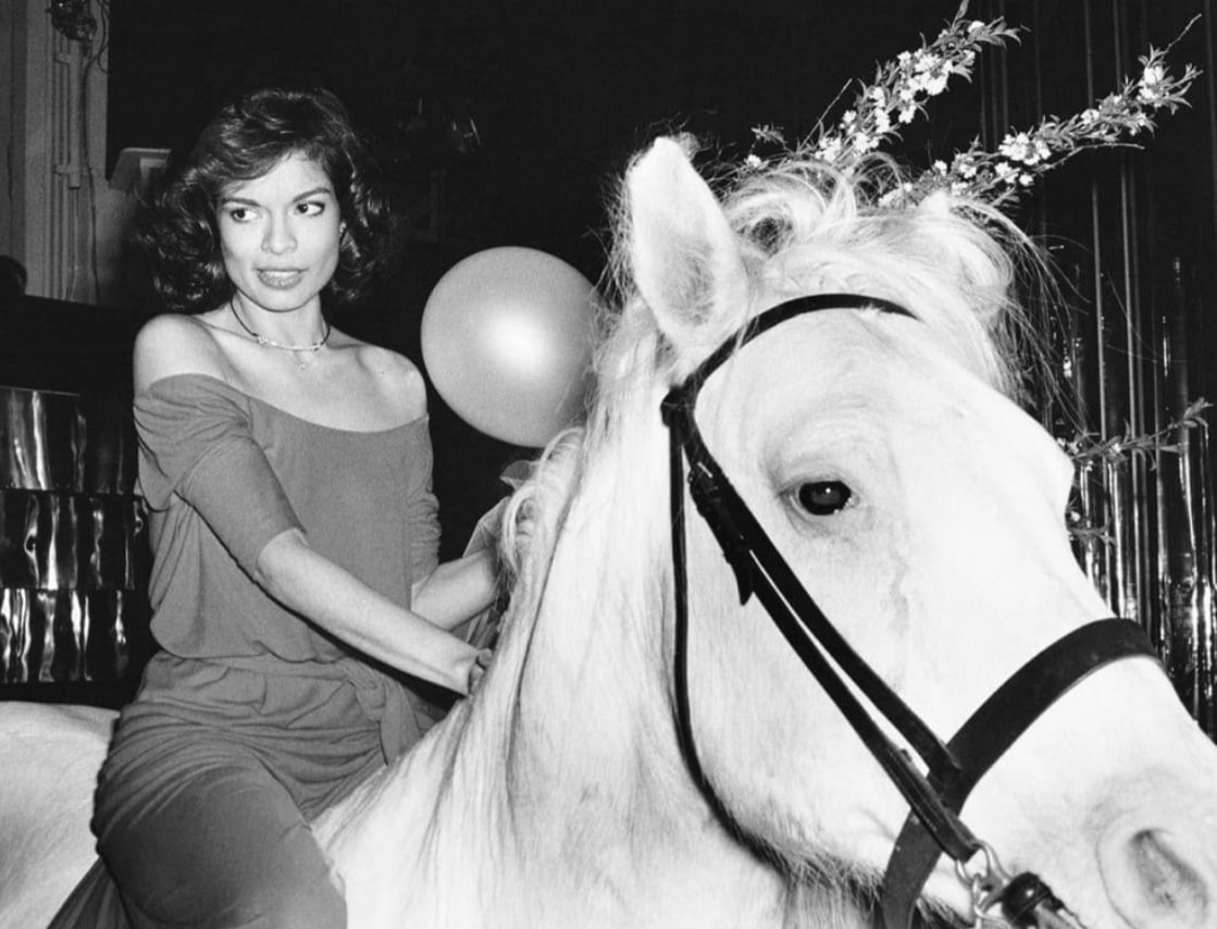 A woman in an off-the-shoulder dress sits on a white horse decorated with flowers, indoors at what appears to be a festive event with a balloon in the background. The image is in black and white.