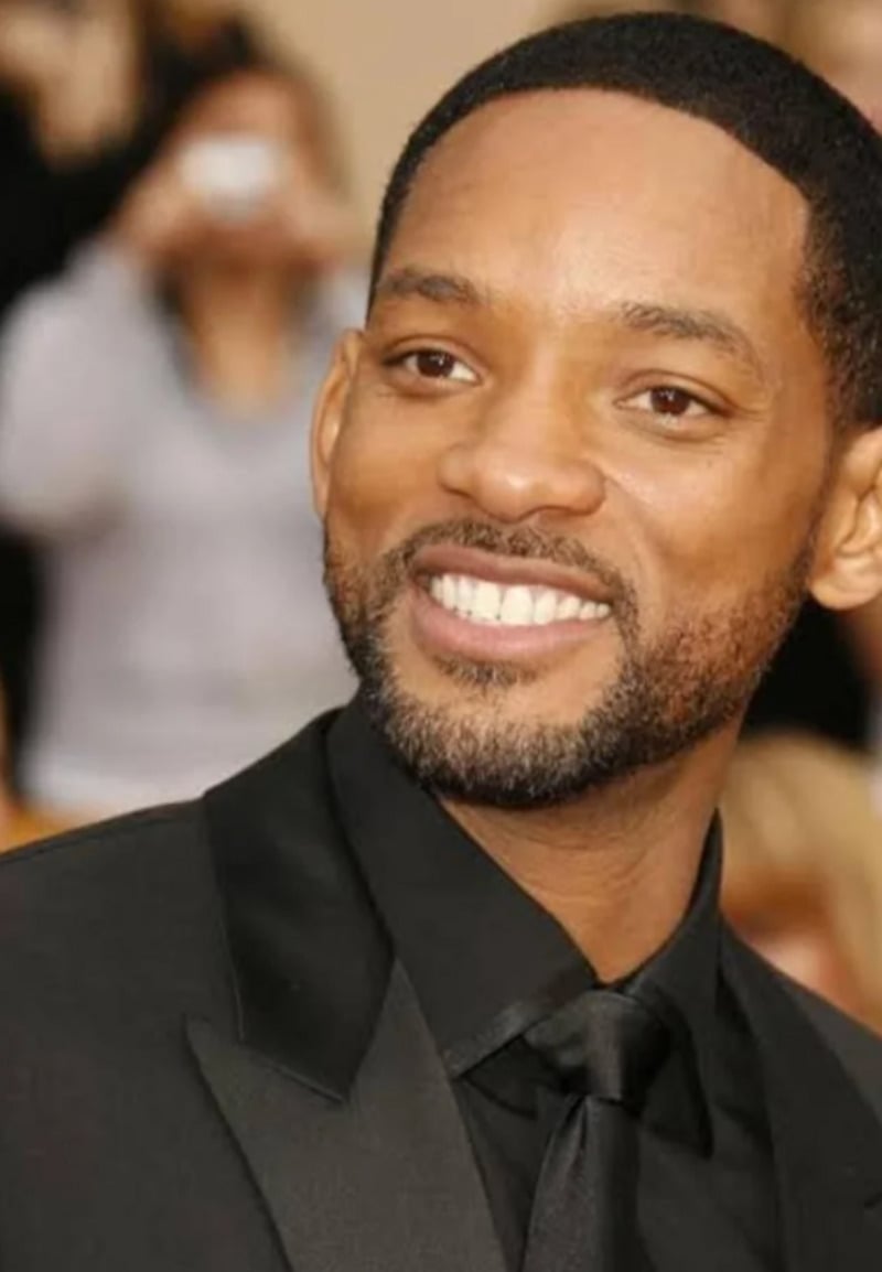 A man in a black suit and tie is smiling at an event. The background is out of focus, showing people and a beige setting.