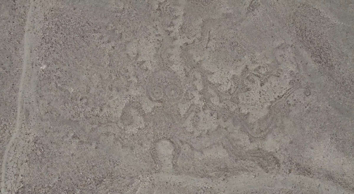 Aerial view of the Nazca Lines, showing a large, intricate geoglyph etched into the desert ground, resembling a stylized figure or abstract shape with lines and curves. The surrounding terrain is dry and sandy.