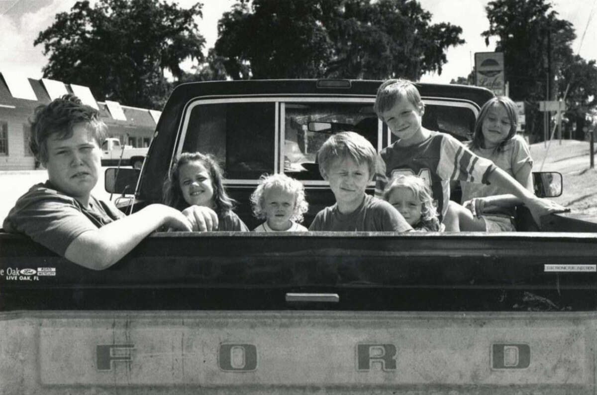 Seven children are sitting in the bed of an old Ford pickup truck, parked on a street. The kids are looking at the camera, some smiling, with trees and buildings visible in the background.