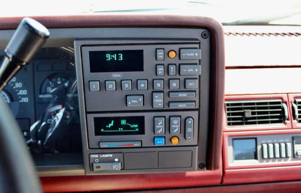 Close-up of an old car dashboard showing a digital clock, audio system controls, air conditioning buttons, and part of the speedometer, all set in a maroon interior.