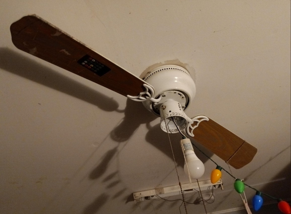 A ceiling fan with three wooden blades, a white light socket containing a light bulb, and colorful string lights hanging nearby. The fan has a pull chain and is mounted on a beige ceiling.