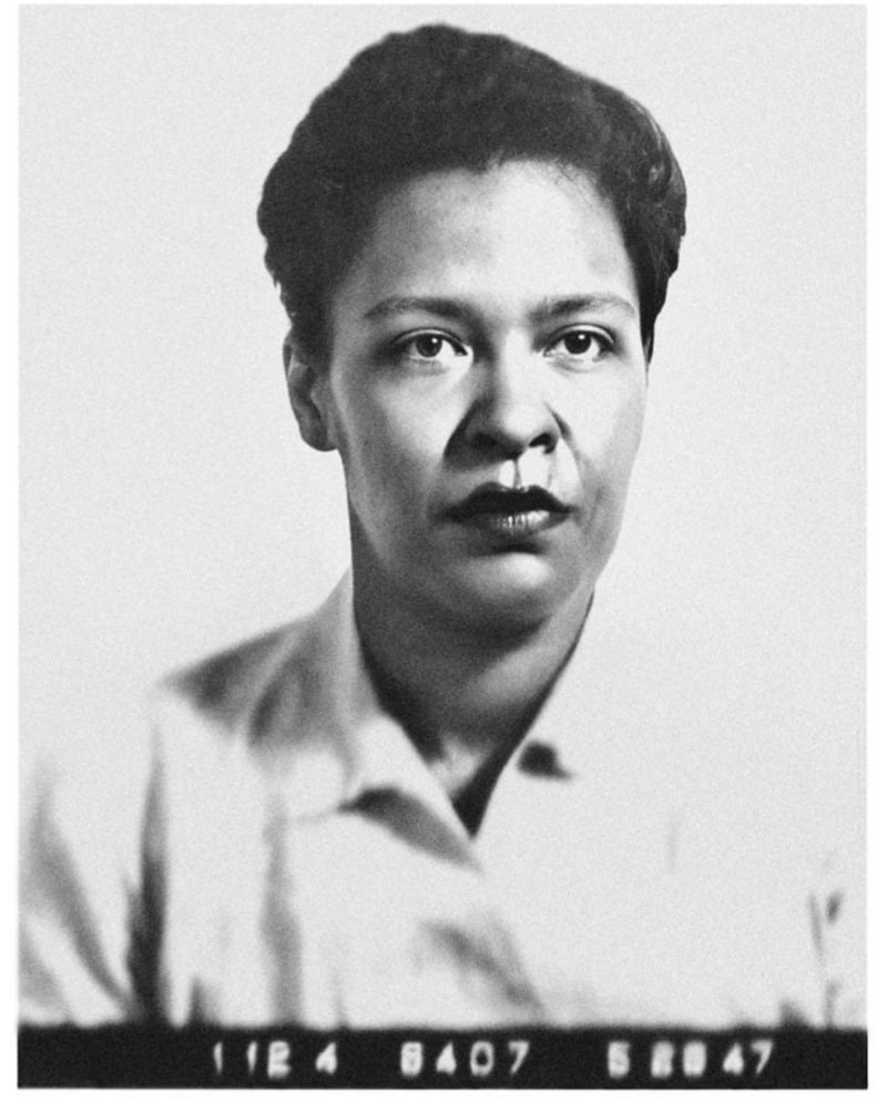 Black and white portrait of a woman with short, curly hair, wearing a collared shirt. She is looking directly at the camera with a neutral expression. The bottom of the image shows a series of numbers.