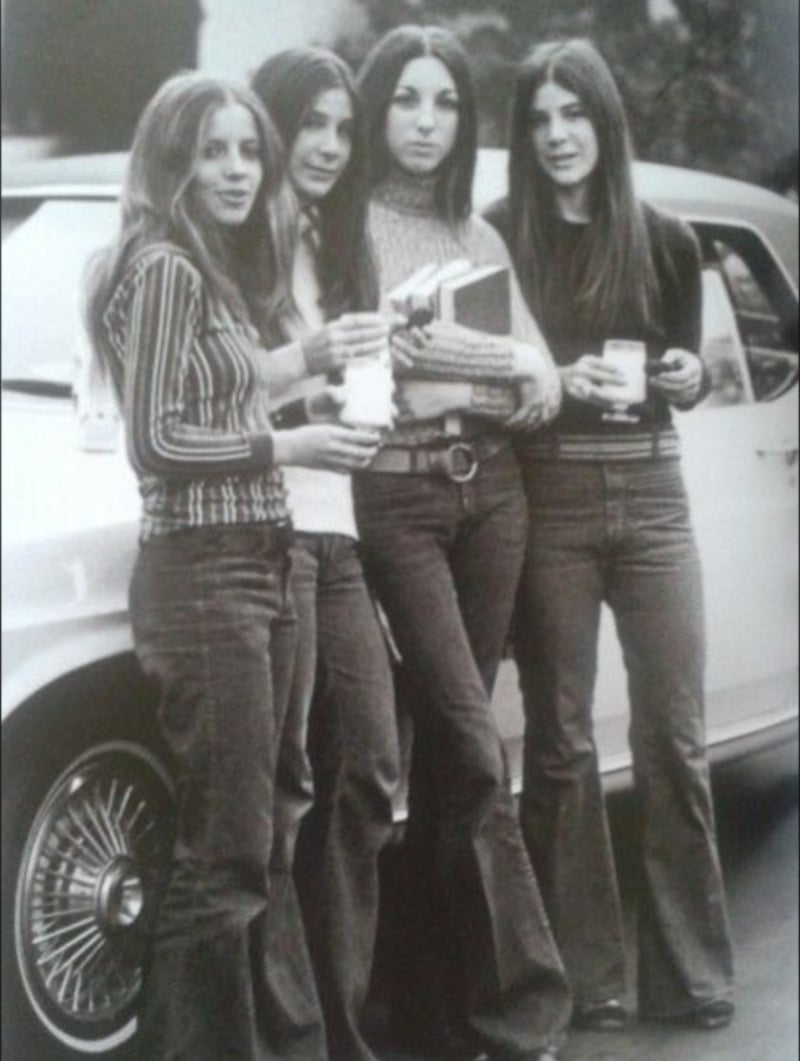 Four young women in flared jeans and long-sleeve tops stand in front of a car, holding drinks and books, posing and smiling for the camera. The image appears to be from the 1970s.