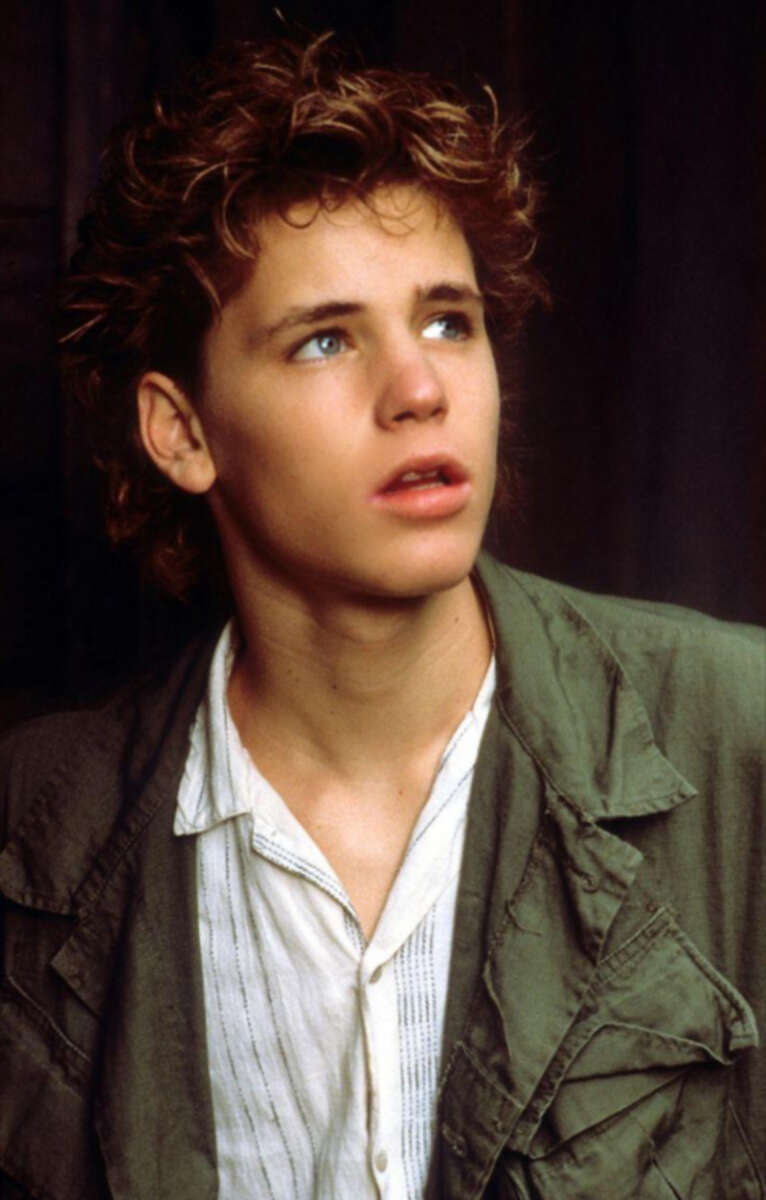 A young person with curly brown hair and blue eyes looks upward with a thoughtful expression. They are wearing a light-colored shirt and an olive green jacket.