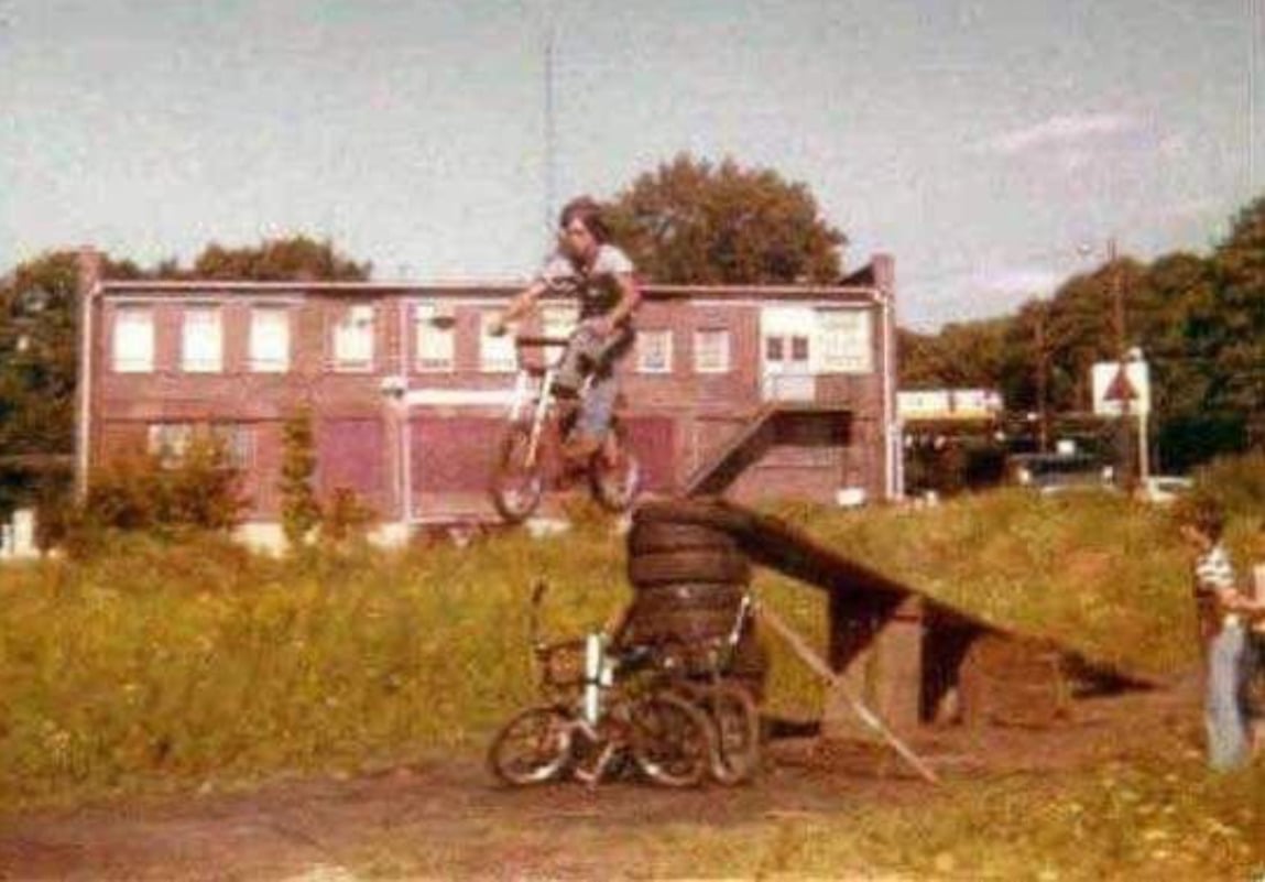 A person on a bicycle jumps off a ramp made of wood and stacked tires, soaring above two parked bikes, with a brick building and trees in the background on a sunny day.